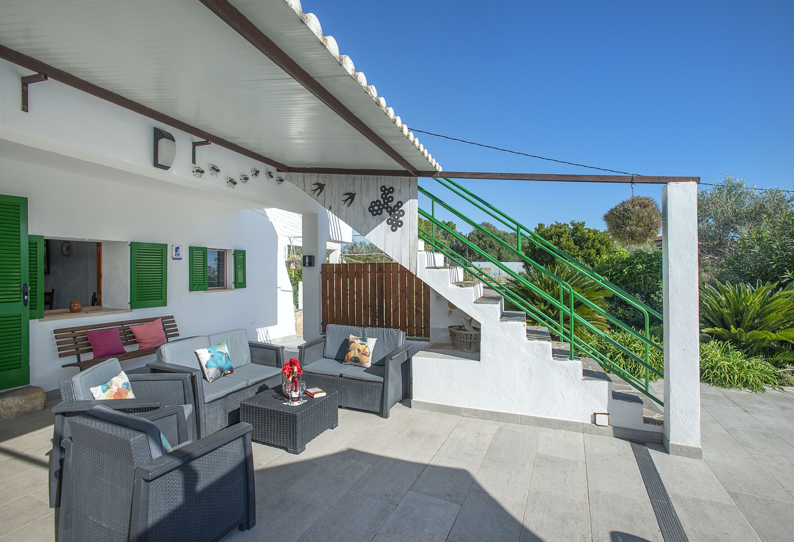 A covered outdoor seating area with wicker furniture, a staircase, and greenery in the background.
