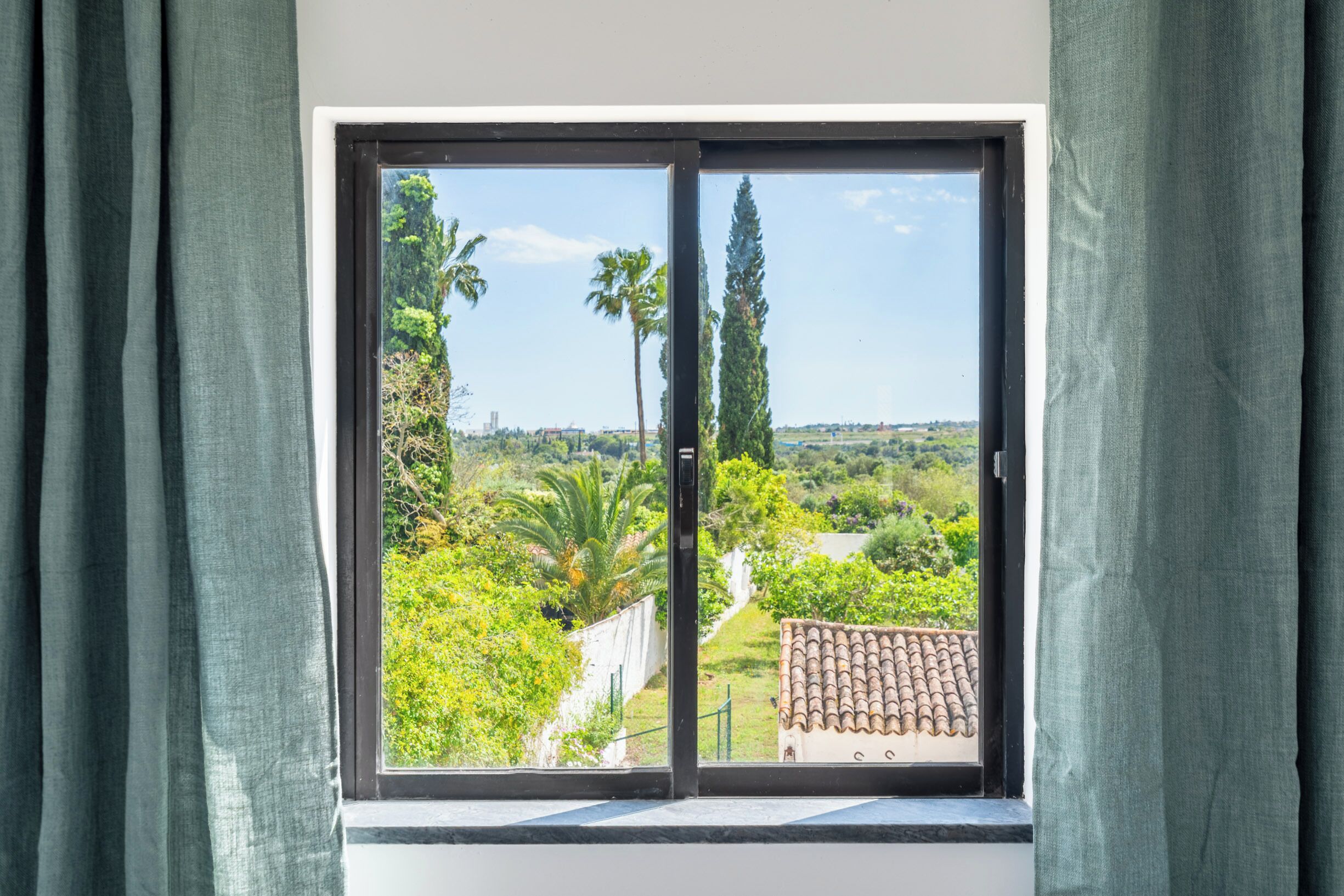 A window with a view of a green landscape, including trees and a building with a tiled roof.