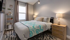 A bedroom with a bed, bedside lamps, a nightstand, a window with curtains, a TV, and a shelf with decorative items.