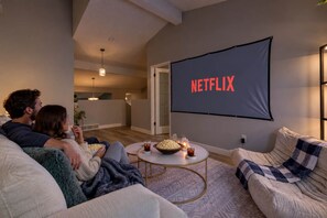 Una sala de estar con una pantalla grande que muestra 'NETFLIX' y un rincón de descanso acogedor con una mesita de centro.