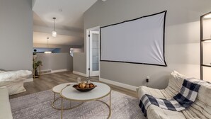 A modern living room with a white sofa, a coffee table with a bowl of fruits, a patterned rug, and a large blank projection screen on the wall.