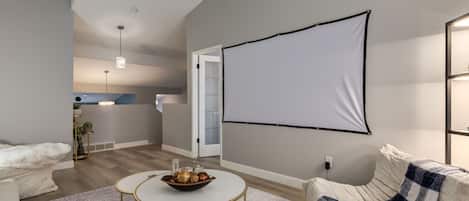 A modern living room with a white sofa, a coffee table with a bowl of fruits, a patterned rug, and a large blank projection screen on the wall.