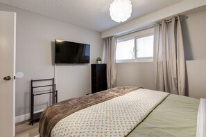Een slaapkamer met een bed, een aan de muur bevestigde televisie, een raam met gordijnen en een kleine plant op een kast.