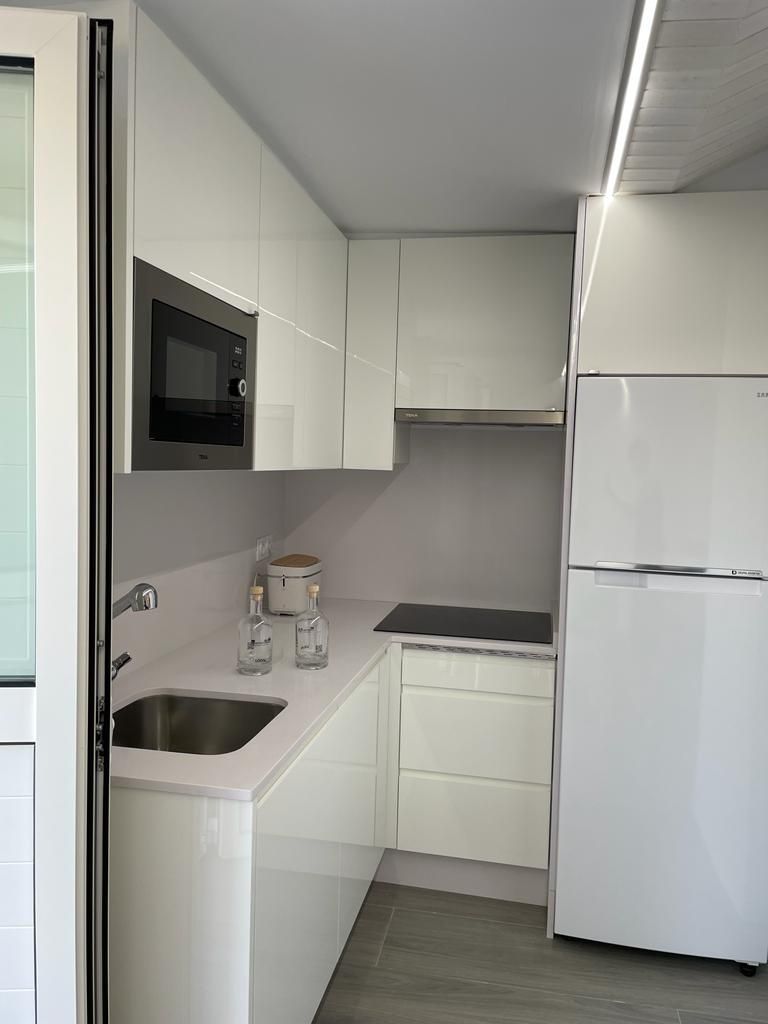 Une cuisine compacte avec des meubles blancs, un four à micro-ondes encastré, un évier en acier inoxydable et un réfrigérateur blanc.