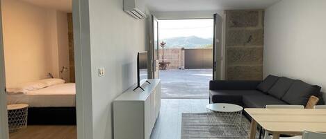 Un moderno salón con sofá, mesa de comedor y televisor. Hay un balcón con vistas a las montañas.