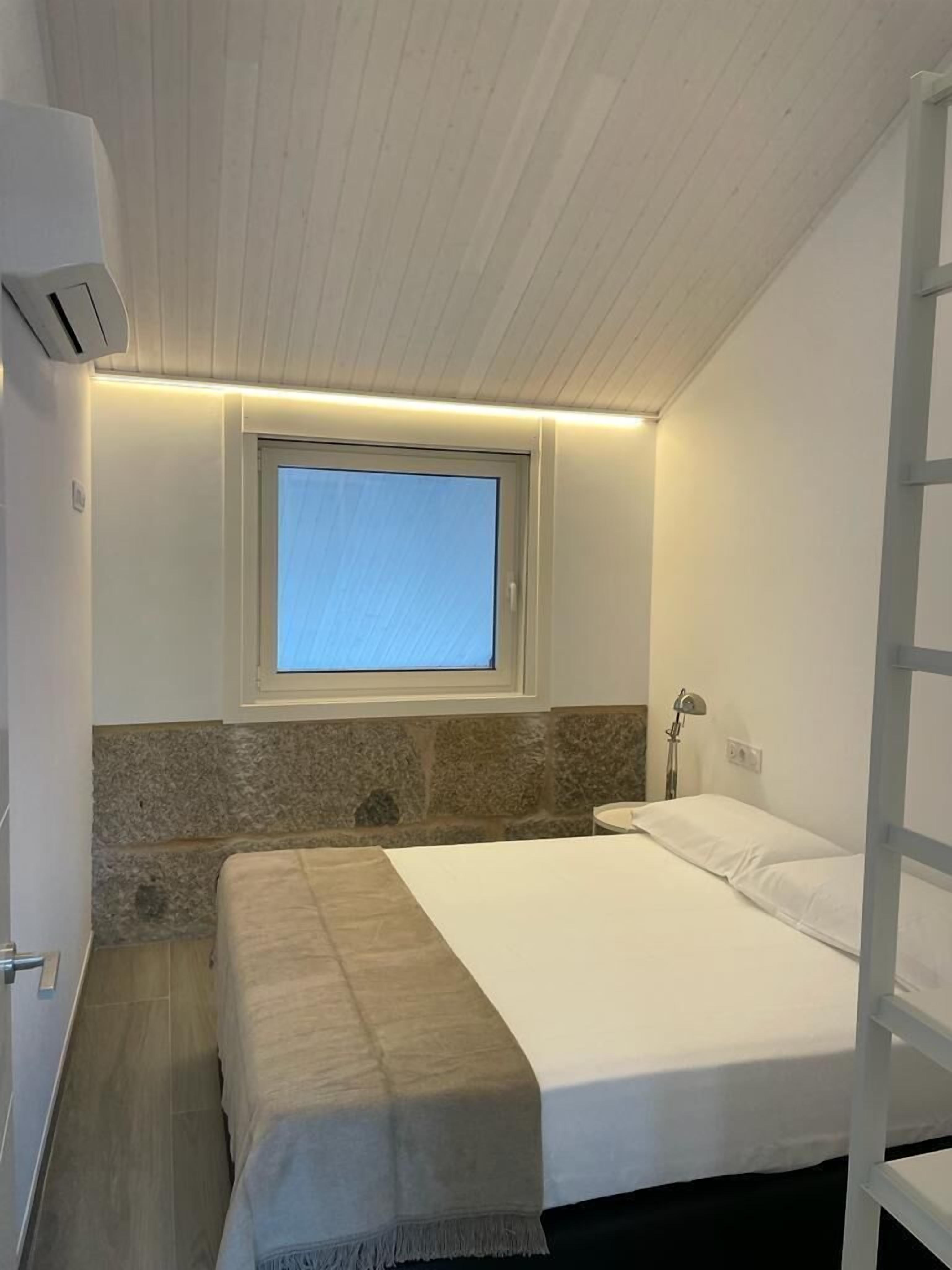 Un dormitorio con cama, ventana, pared de piedra, mesita de noche y lámpara.