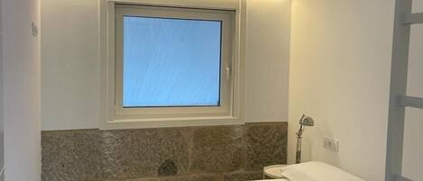 Un dormitorio con cama, ventana, pared de piedra, mesita de noche y lámpara.
