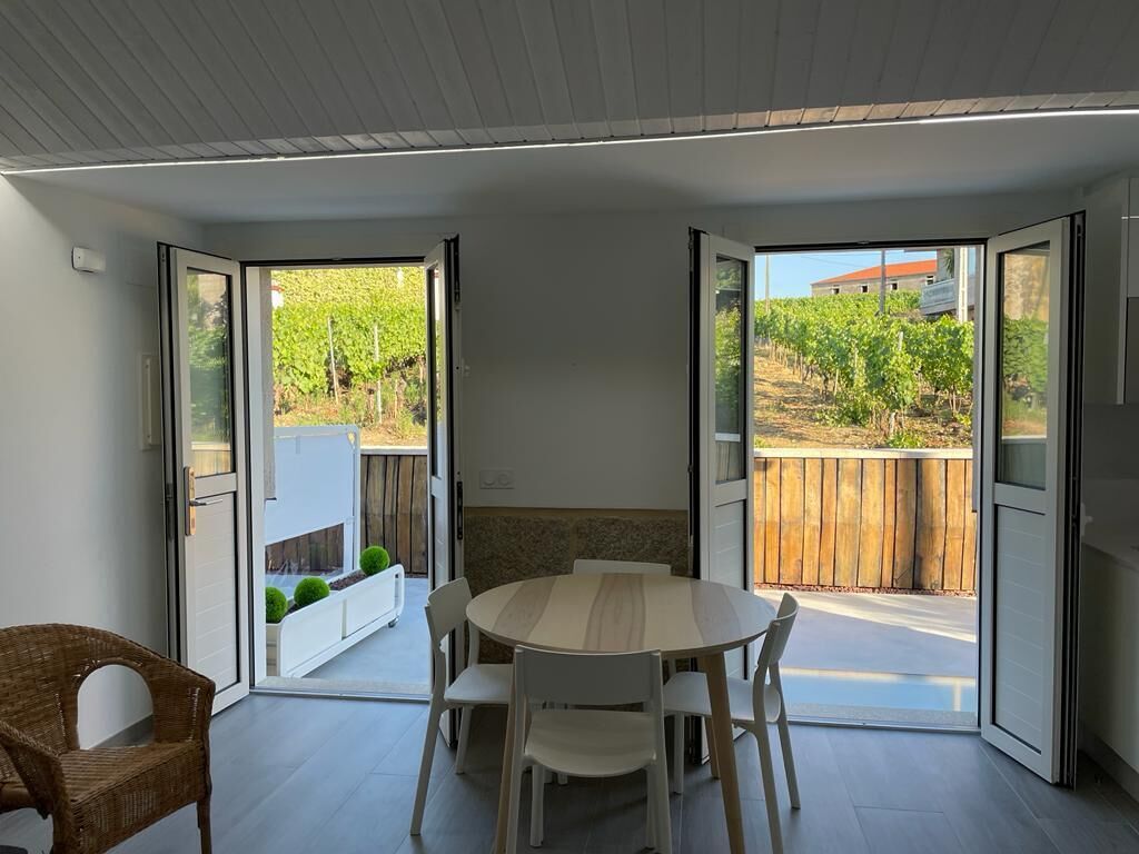 A modern dining area with a round table and chairs, large sliding glass doors leading to a garden with a wooden fence and greenery.