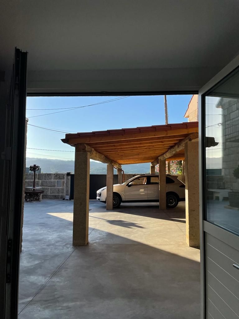 Un espace extérieur abrité, avec une voiture garée en dessous, une table avec des chaises, et une vue sur les montagnes en arrière-plan.