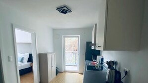 Een kleine keuken met een spoelbak, waterkoker en keukenkastjes. Een deur leidt naar een slaapkamer met een bed en een raam.