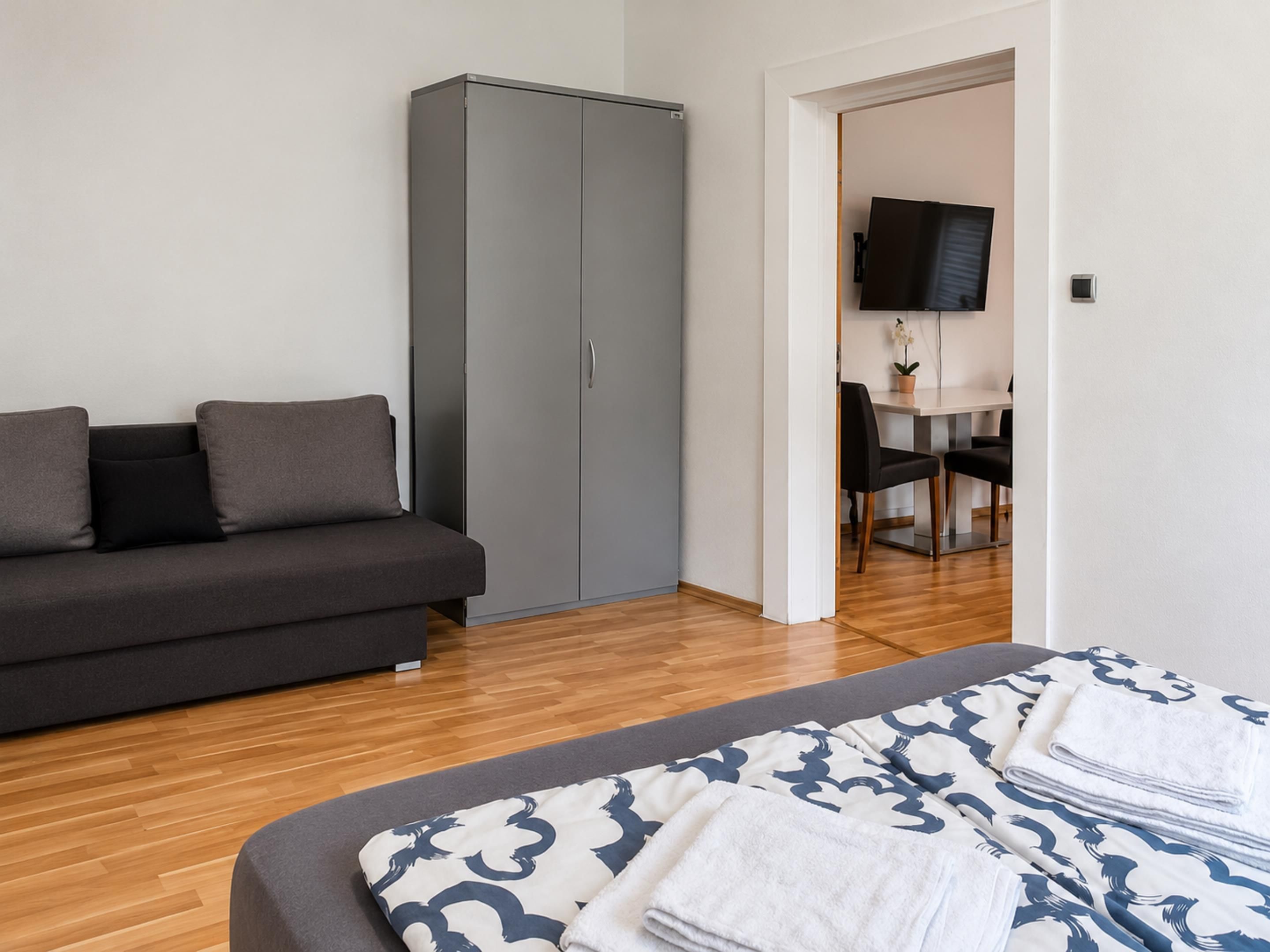 A modern bedroom with a grey sofa, a wardrobe, a bed with patterned bedding, and a television in the adjacent room.