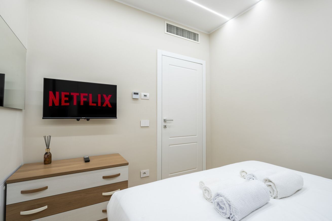 A hotel room with a bed, a TV mounted on the wall displaying "NETFLIX," a wooden dresser, and a white door.