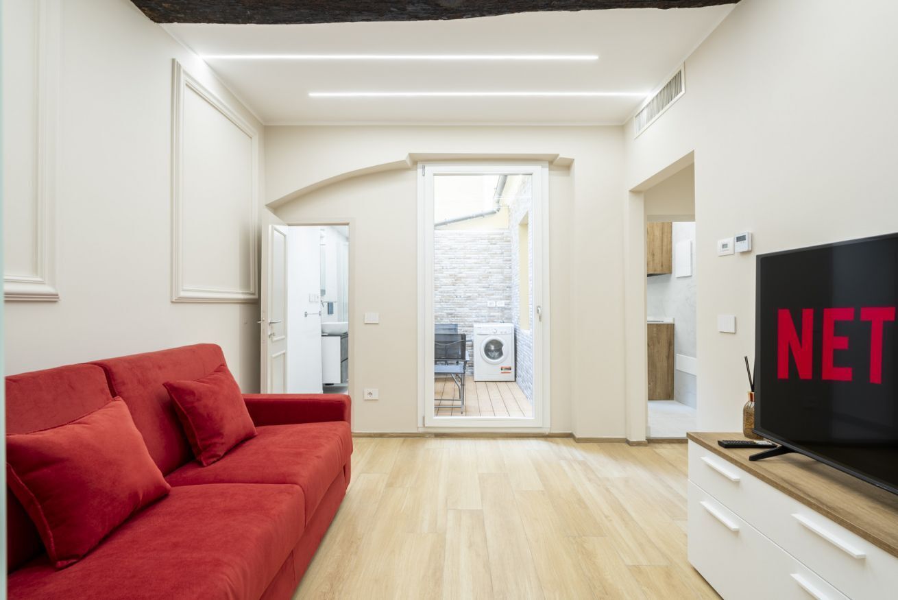Un salon moderne avec un canapé rouge, un téléviseur et une vue sur la cuisine et l’espace buanderie.