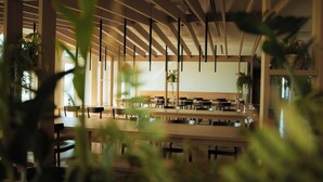 Un vaste espace intérieur doté de poutres et de tables en bois, agrémenté de grandes fenêtres et de plantes en pot.