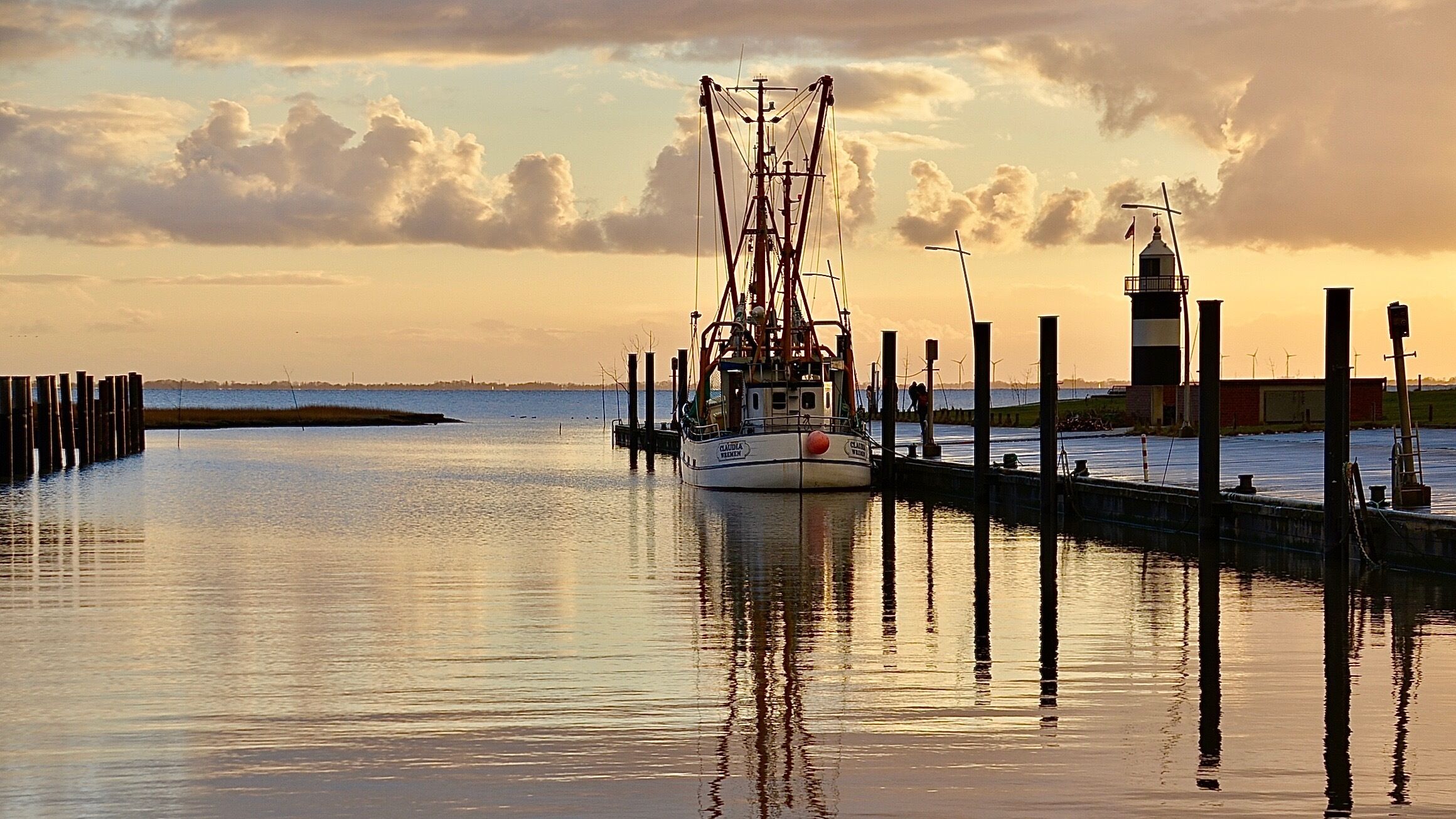A calm harbor with a fishing boat docked at a pier, a lighthouse, and a calm sea reflecting the sky.
