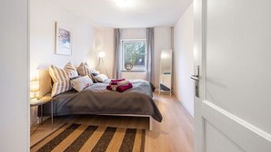 A bedroom with a bed, a bedside table with a lamp, a window with curtains, and a door. - Apartment 'Horsti' with Wi-Fi (Timmendorfer Strand)