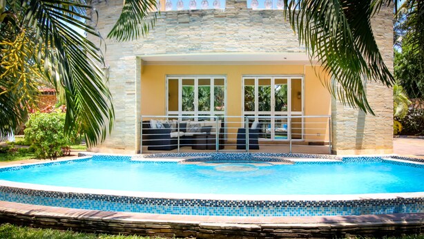 Une piscine avec un bord carrelé, un espace détente couvert comprenant un canapé et des fauteuils, et un mur de pierre en arrière-plan.