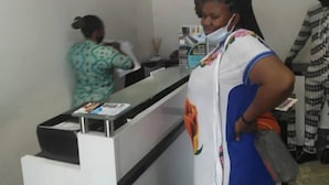 Un área de recepción con mostrador, una mujer vestida con estampado y otra mujer con un vestido azul y mascarilla facial.
