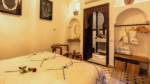 A hotel room with a bed, a chair, a vanity with a mirror, a shelf with toiletries, and a clothing rack with towels. - Elegant Riad Musée: Authentic Marrakech Charm in the Medina (marrakech)