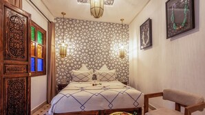 A bedroom with a patterned wallpaper, a bed with a geometric bedspread, a wooden chair, and a small table with a decorative bowl. - Elegant Riad Musée: Authentic Marrakech Charm in the Medina (marrakech)