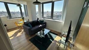 Un salon moderne avec un canapé gris, des coussins noirs et turquoise, une table basse en verre et une vue sur la ville grâce à de grandes fenêtres.