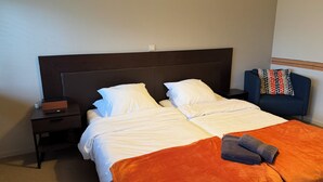 Ein Doppelbett mit weißen Laken, einer orangefarbenen Decke und einem blauen Handtuch. Ein Nachttisch mit einem Buch und einer Lampe steht daneben. Ein blauer Stuhl mit einem gemusterten Kissen lehnt an der Wand.