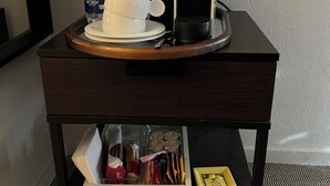Ein kleiner Holznachttisch mit Kaffeemaschine, einer Wasserflasche und einer Tasse darauf. Darunter befindet sich eine Schublade mit verschiedenen Gegenständen.