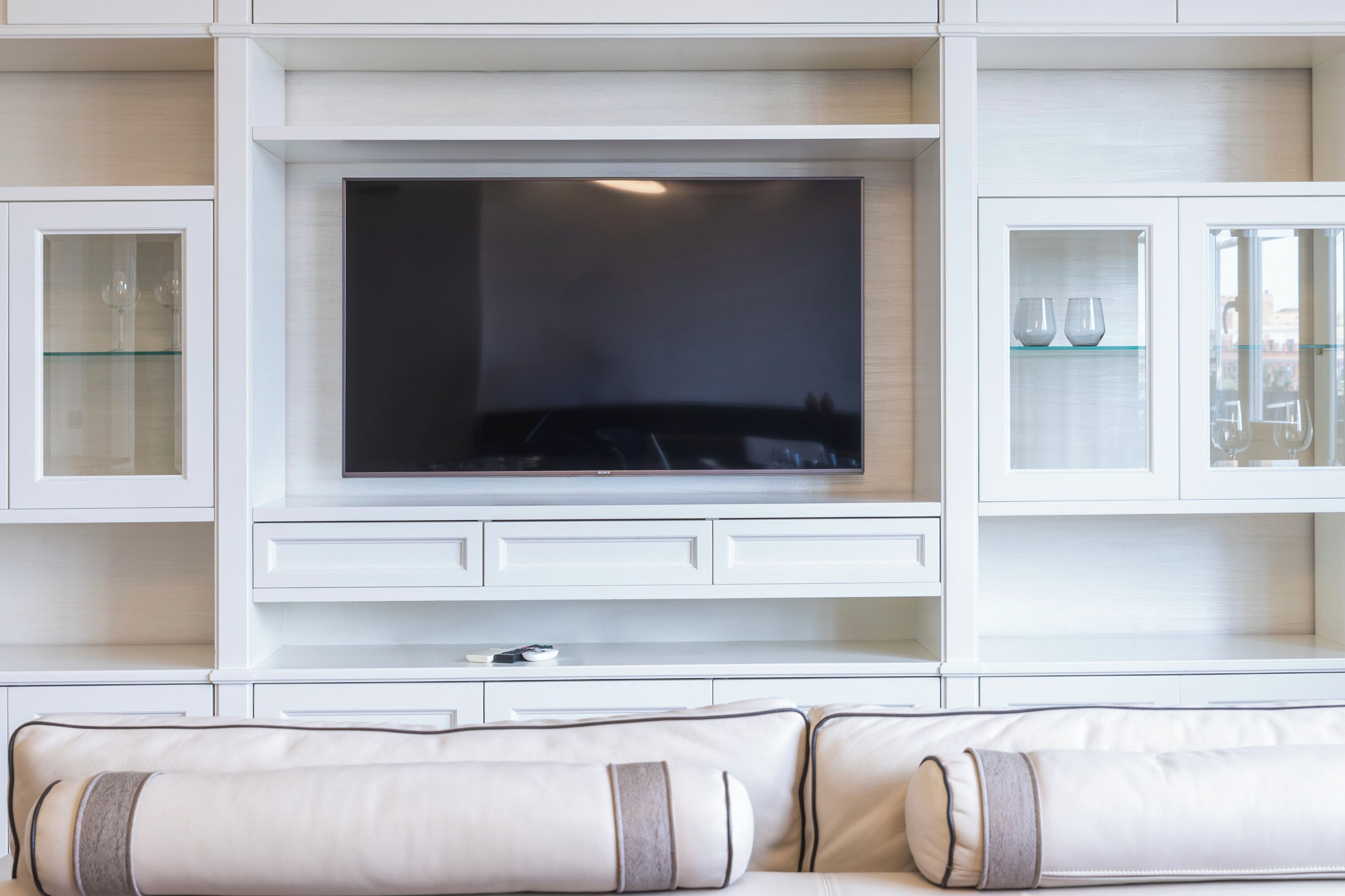 Ein modernes Wohnzimmer mit einem Flachbildfernseher an einem weißen Schrank, einem Glastischchen mit Weingläsern und einem beigefarbenen Sofa mit gemusterten Kissen.
