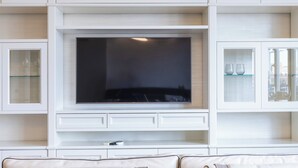 Un salon moderne avec un téléviseur à écran plat fixé sur un meuble blanc, une étagère en verre où sont posés des verres à vin, et un canapé beige agrémenté de coussins à motifs.