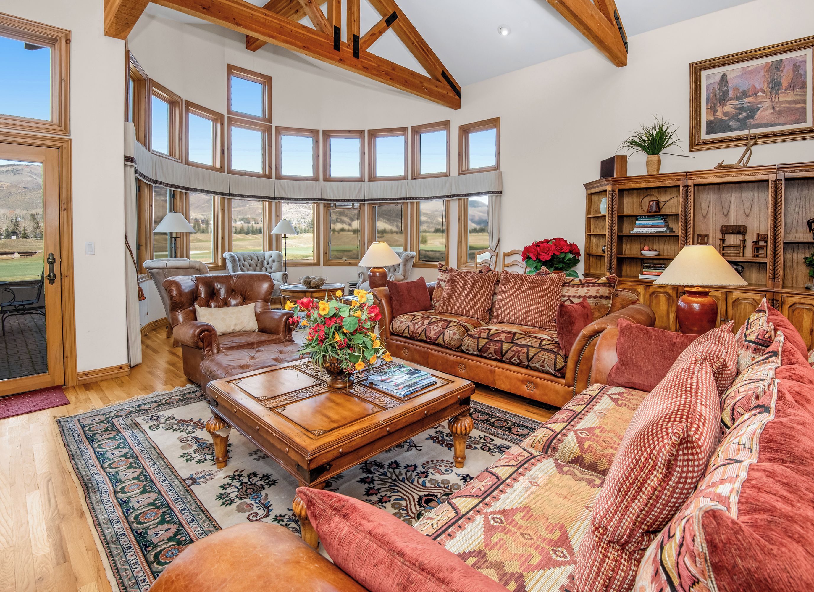 A spacious living room with a large window, leather sofas, a wooden coffee table, and a bookshelf.