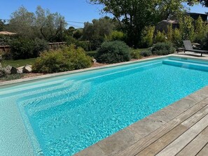 Ein rechteckiger Swimmingpool mit klarem, blauem Wasser, umgeben von einer Holzterrasse und verschiedenen Sträuchern und Bäumen.
