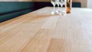 Een houten tafel met een fles en glazen, een klok aan de muur, en een groene, beklede bank.