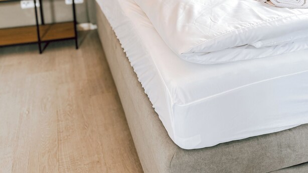 Een netjes opgemaakt bed met witte lakens en een opgerolde handdoek, een houten ladder en een lichtgekleurde vloer.