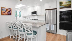Una cocina moderna con armarios blancos, un refrigerador de acero inoxidable y un horno doble. Hay taburetes de bar con cojines color turquesa y un cuadro en la pared.