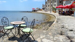A coastal area with stone tables and chairs, a stone building, and red umbrellas. - Holiday home with garden and BBQ in Umag (Umag)