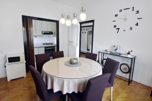 Een eetruimte met een ronde tafel, stoelen, een klok en een keuken op de achtergrond.