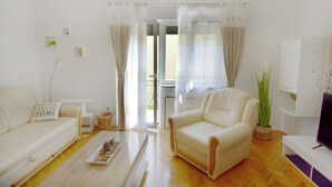 En moderne stue med sofa, lænestol og sofabord. Der er et fjernsyn på et stativ og en plante i en kurv.