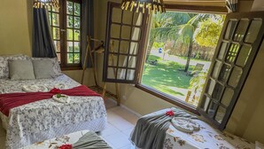 Ein Zimmer mit zwei Betten, jedes mit floralem Bettbezug und einer roten Rose. Es gibt große Fenster mit Holzläden, eine hängende Leuchte und einen kleinen Tisch mit einer Pflanze.