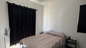 Un dormitorio con cama, un ventilador, una mesita con una planta y una ventana con cortinas.
