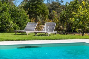 Piscina com duas espreguiçadeiras e área gramada com  árvores.