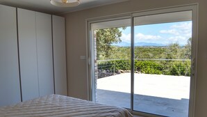 Et soveværelse med en seng og en skydedør, der fører ud til en balkon med udsigt over grøn vegetation og bjerge.