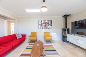 Uma sala de estar moderna com um sofá vermelho, duas poltronas amarelas, uma mesa de centro de madeira, um tapete listrado e colorido, um quadro na parede, uma televisão e uma lareira.