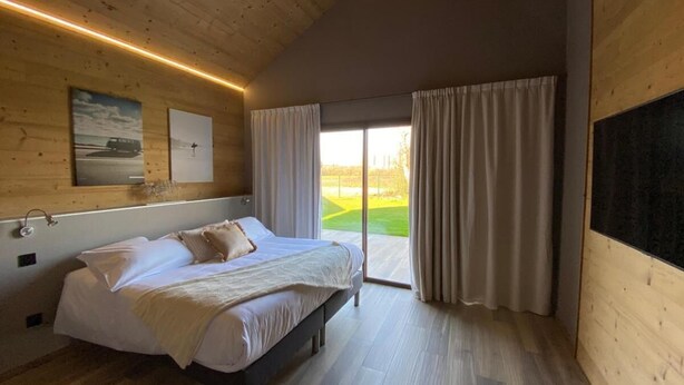 A bedroom with a bed, wooden walls, a large window with curtains, and a view of a green landscape. - WORLD HOTEL (Saint-Vulbas)