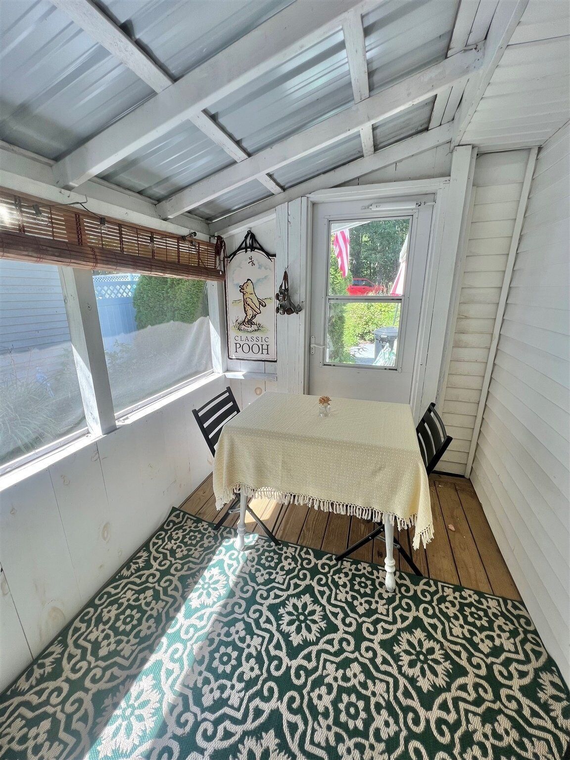A small, enclosed porch with a table and chairs, a hanging sign, and a window with a view of the outside.