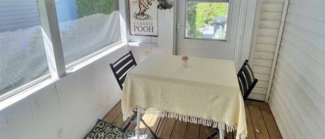 A small, enclosed porch with a table and chairs, a hanging sign, and a window with a view of the outside.