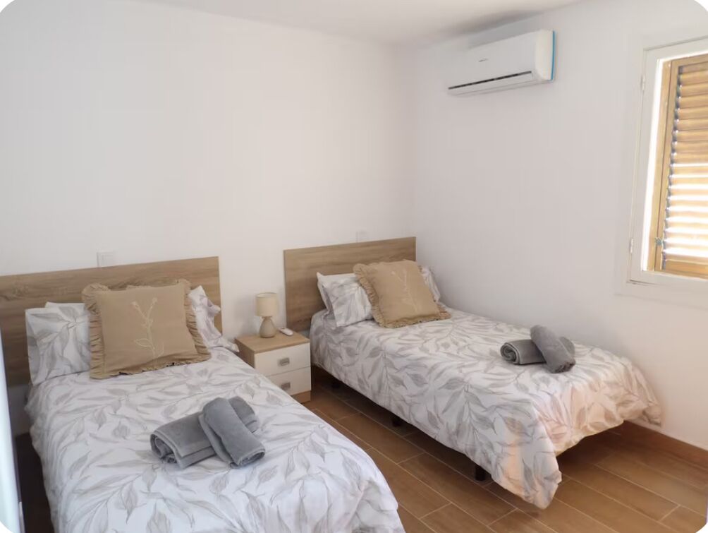 Een slaapkamer met twee bedden, elk voorzien van een houten hoofdbord, nachtkastje en een raam met jaloezieën.