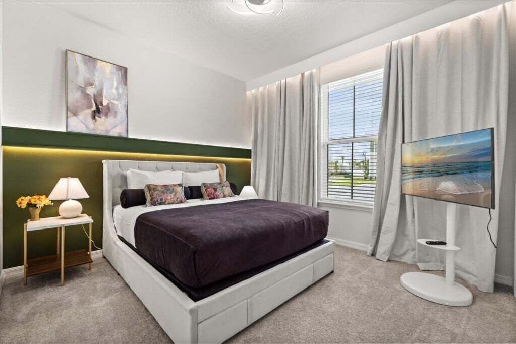 A modern bedroom with a large bed, a bedside table with a lamp, a painting on the wall, and a window with curtains.