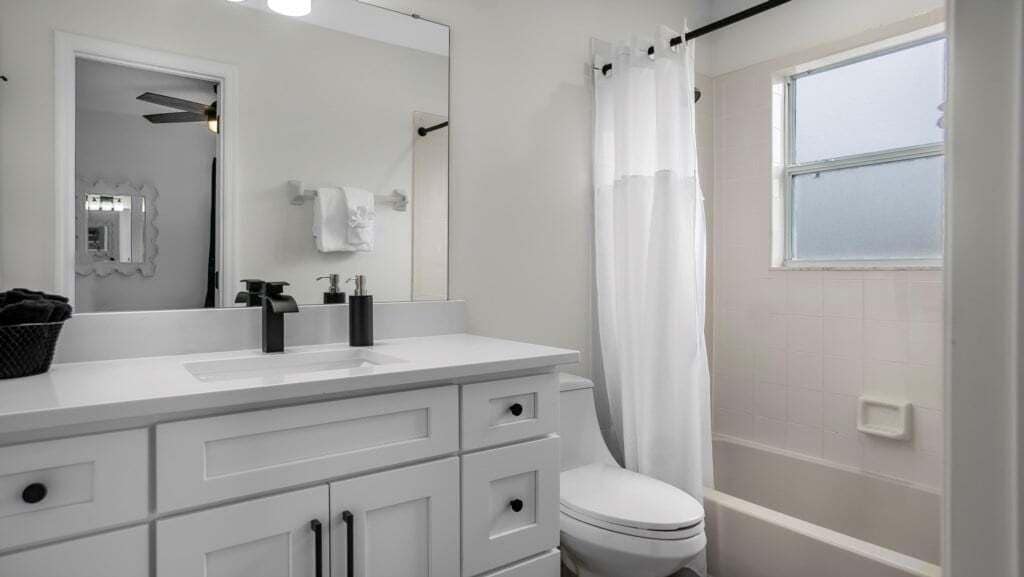 Un baño moderno con un tocador blanco, inodoro, ducha con cortina y ventana que proporciona luz natural.