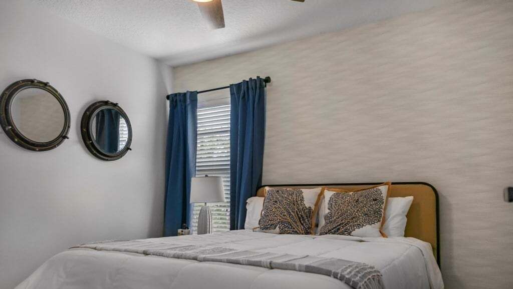 A bedroom with a bed, two wall-mounted mirrors, a window with blue curtains, and a light fixture on the ceiling.