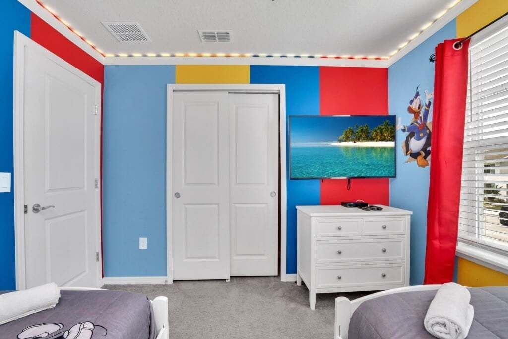 A room with two beds, a white dresser, a TV on the wall, and a red curtain.